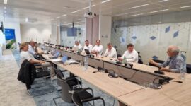 Wooden U‑shaped conference table with several people in a meeting room, laptops open and microphones ready for discussion.