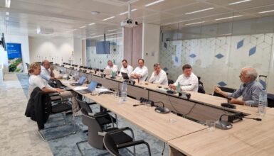 Wooden U‑shaped conference table with several people in a meeting room, laptops open and microphones ready for discussion.
