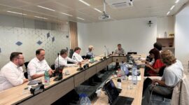 Nine professionals sit around a long curved conference table in a modern office, laptops and papers in use, with water bottles and coffee cups nearby and a ceiling projector overhead.