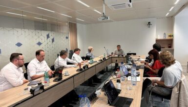 Nine professionals sit around a long curved conference table in a modern office, laptops and papers in use, with water bottles and coffee cups nearby and a ceiling projector overhead.