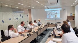 A meeting room with nine people seated around a long conference table, several using laptops, watching a video conference projected on the wall screen at the far end.