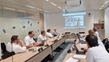 A meeting room with nine people seated around a long conference table, several using laptops, watching a video conference projected on the wall screen at the far end.