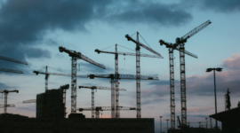 Group of tall construction cranes over a building site at dusk, dark clouds in the sky.