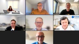 Seven-person video conference grid; participants in offices and homes, with the top-center speaker highlighted in a brown shirt.