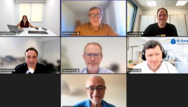 Seven-person video conference grid; participants in offices and homes, with the top-center speaker highlighted in a brown shirt.