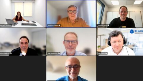 Seven-person video conference grid; participants in offices and homes, with the top-center speaker highlighted in a brown shirt.