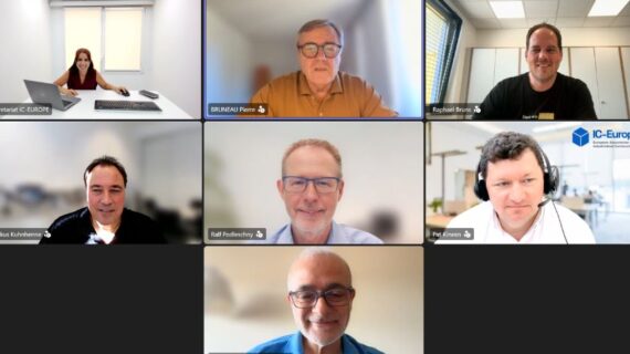 Seven-person video conference grid; participants in offices and homes, with the top-center speaker highlighted in a brown shirt.