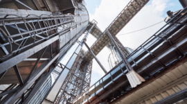 Industrial steel framework with pipes, ladders, and catwalks seen from below against a blue sky in an industrial facility.