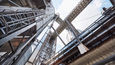 Industrial steel framework with pipes, ladders, and catwalks seen from below against a blue sky in an industrial facility.